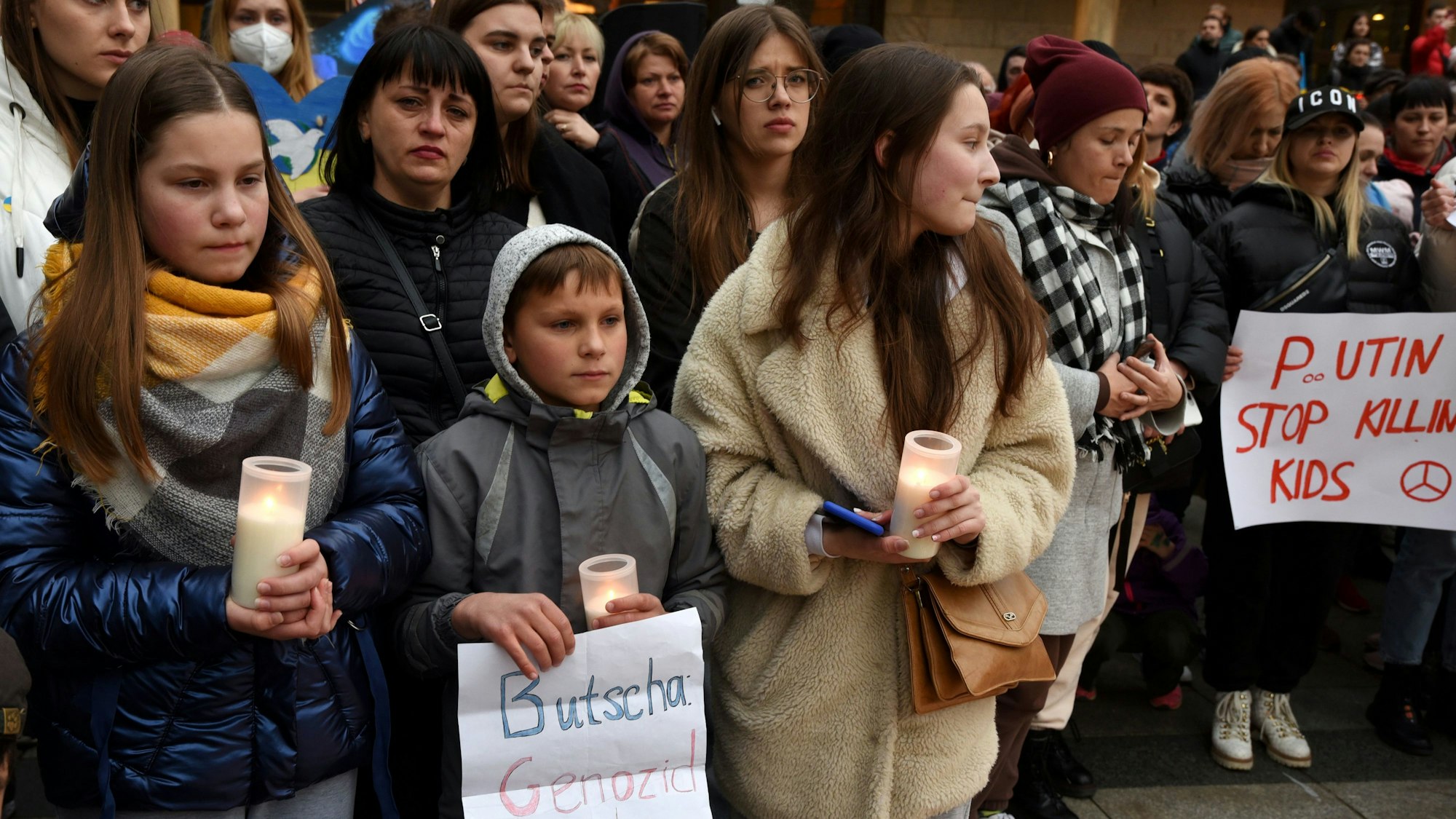 Kinder und Jugendliche halten Kerzen und Plakate, darauf steht unter anderem „Putin, Stop Killing Kids“.