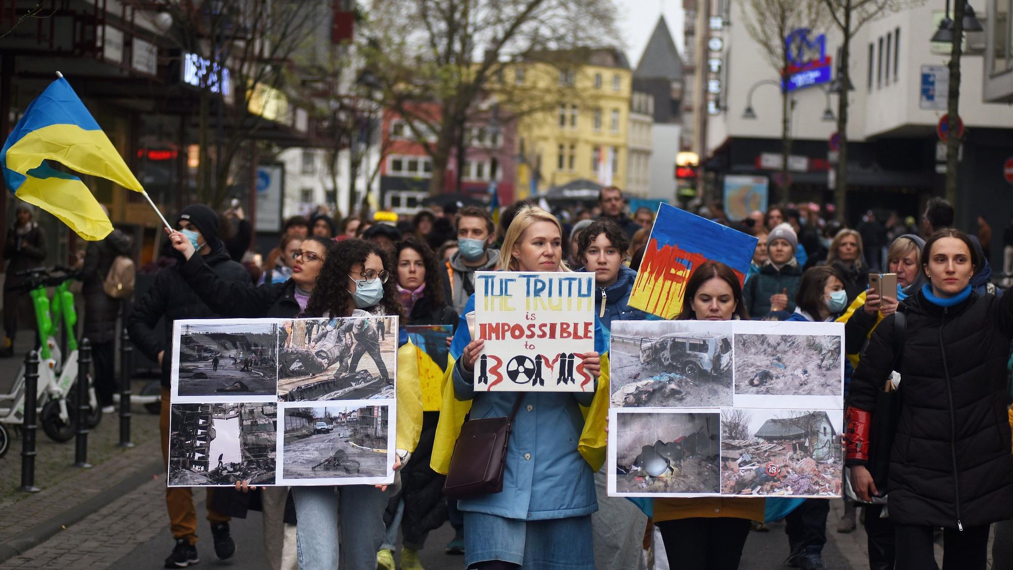 Teilnehmer einer Kundgebung laufen durch eine Straße, sie halten Plakate mit Fotos unter anderem von zerstörten Häusern in der Ukraine in Händen.