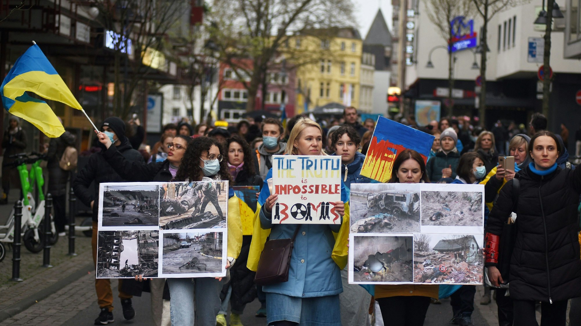 Teilnehmer einer Kundgebung laufen durch eine Straße, sie halten Plakate mit Fotos unter anderem von zerstörten Häusern in der Ukraine in Händen.