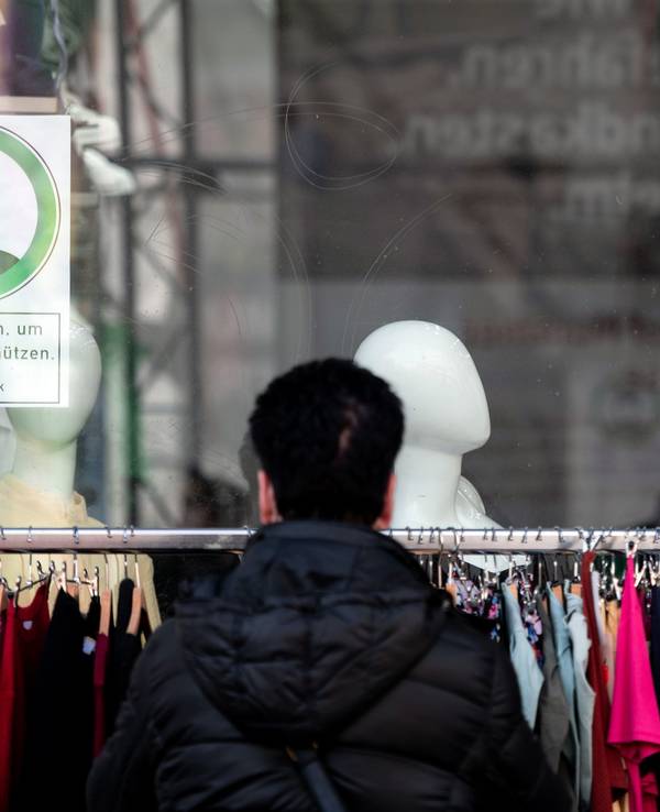 Eine Frau schaut sich Kleidung an einer Stange vor einem Geschäft an, auf einem Schild im Fenster steht "Maske tragen, um andere zu schützen".
