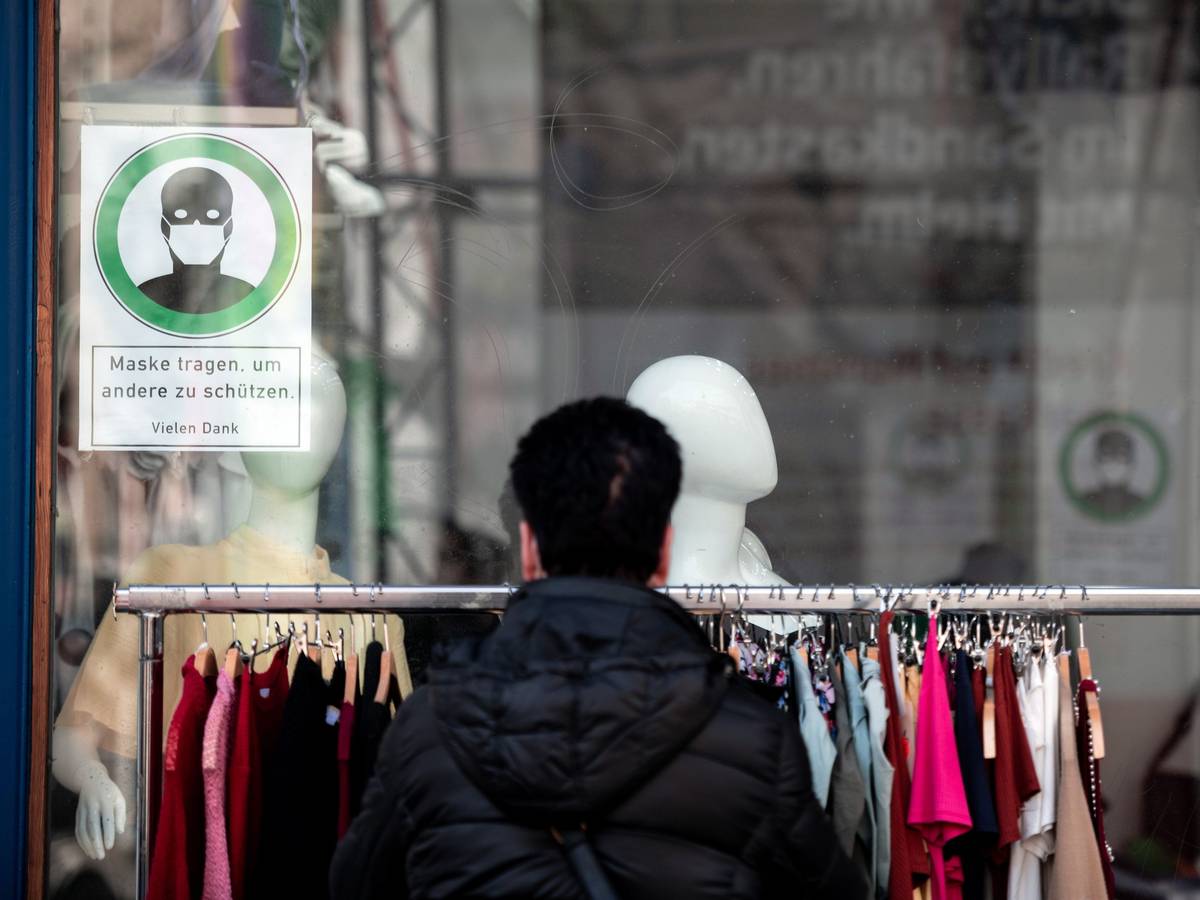 Eine Frau schaut sich Kleidung an einer Stange vor einem Geschäft an, auf einem Schild im Fenster steht "Maske tragen, um andere zu schützen".