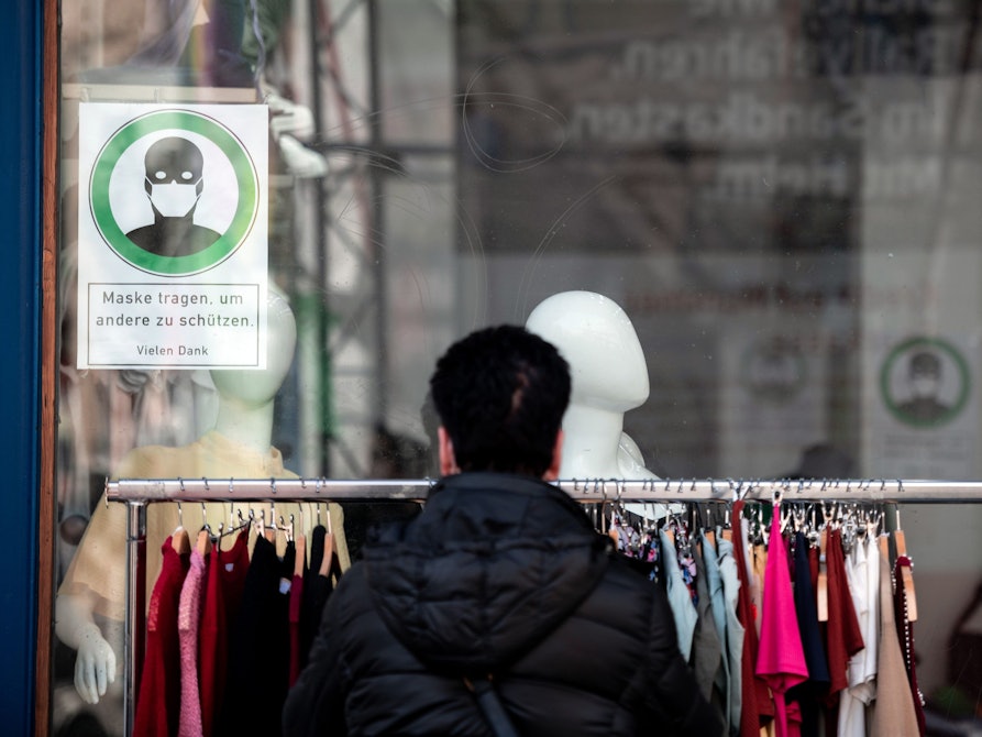 Eine Frau schaut sich Kleidung an einer Stange vor einem Geschäft an, auf einem Schild im Fenster steht "Maske tragen, um andere zu schützen".
