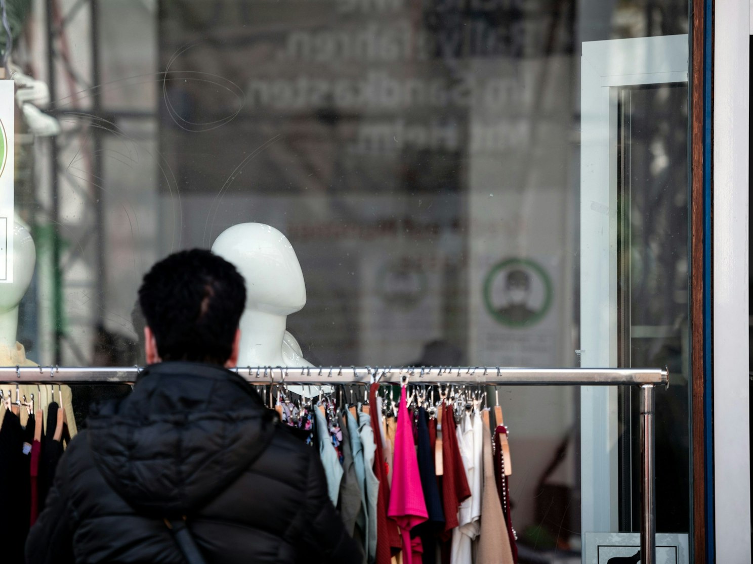Eine Frau schaut sich Kleidung an einer Stange vor einem Geschäft an, auf einem Schild im Fenster steht "Maske tragen, um andere zu schützen".