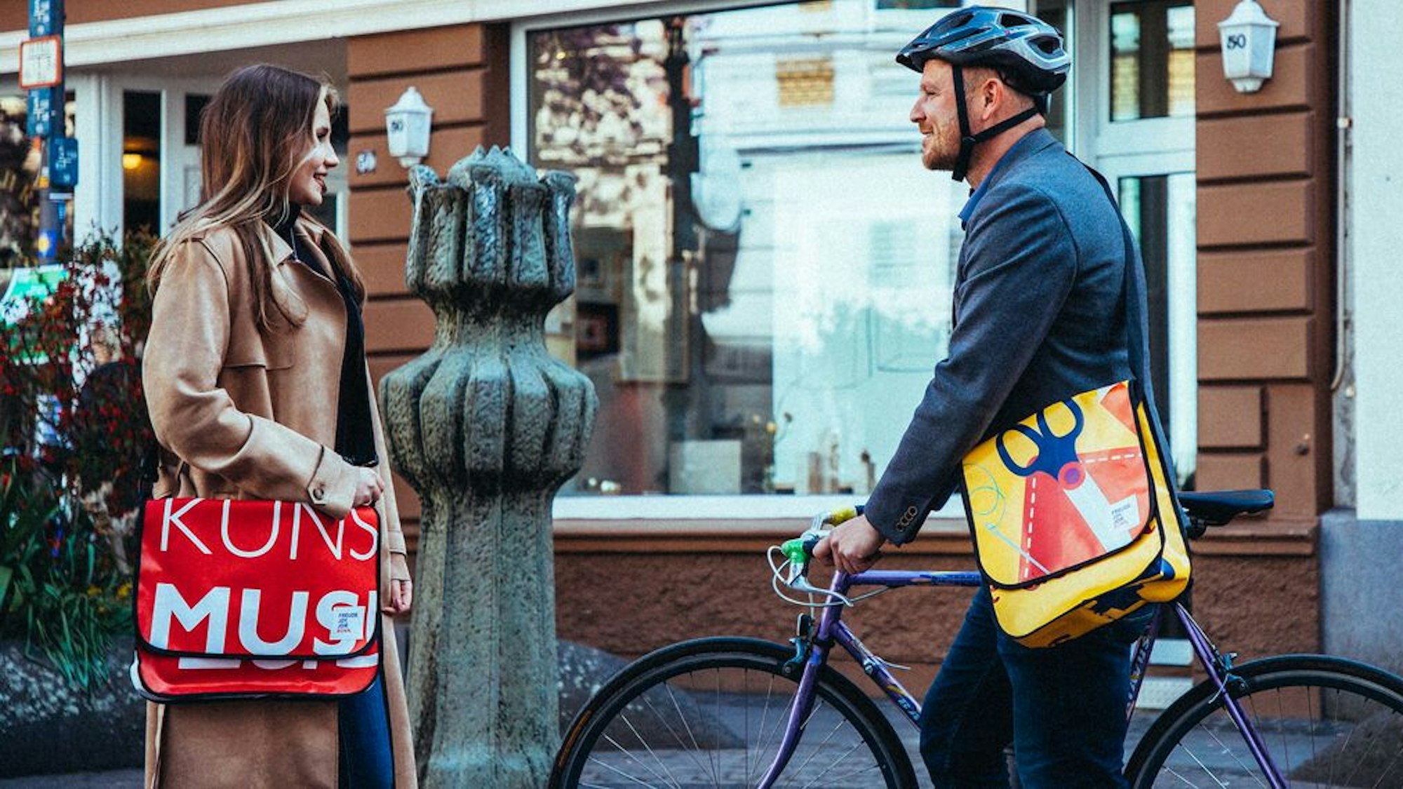Ein Mann und eine Frau tragen Umhängetaschen, auf der einen ist ein Teil des Schriftzugs „Kunstmuseum“ zu erkennen, auf der anderen ist eine Schere zu sehen.