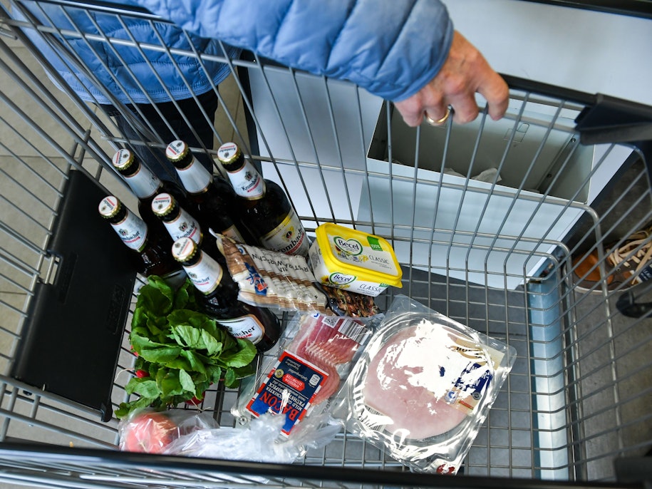 Ein Kunde hat Bier, Wurst, Margarine, Schinken und Salat in einen Einkaufswagen gelegt.