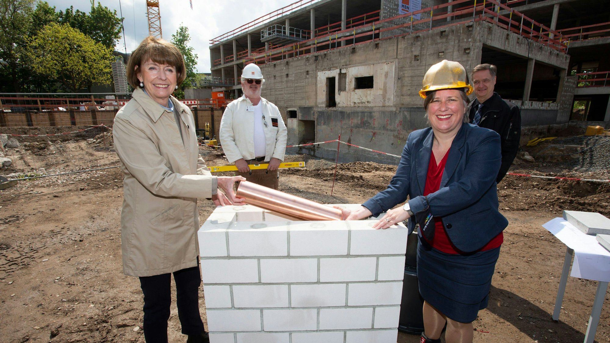 Henriette Reker und drei weitere Personen legen vor einem Gebäude einen Grundstein.