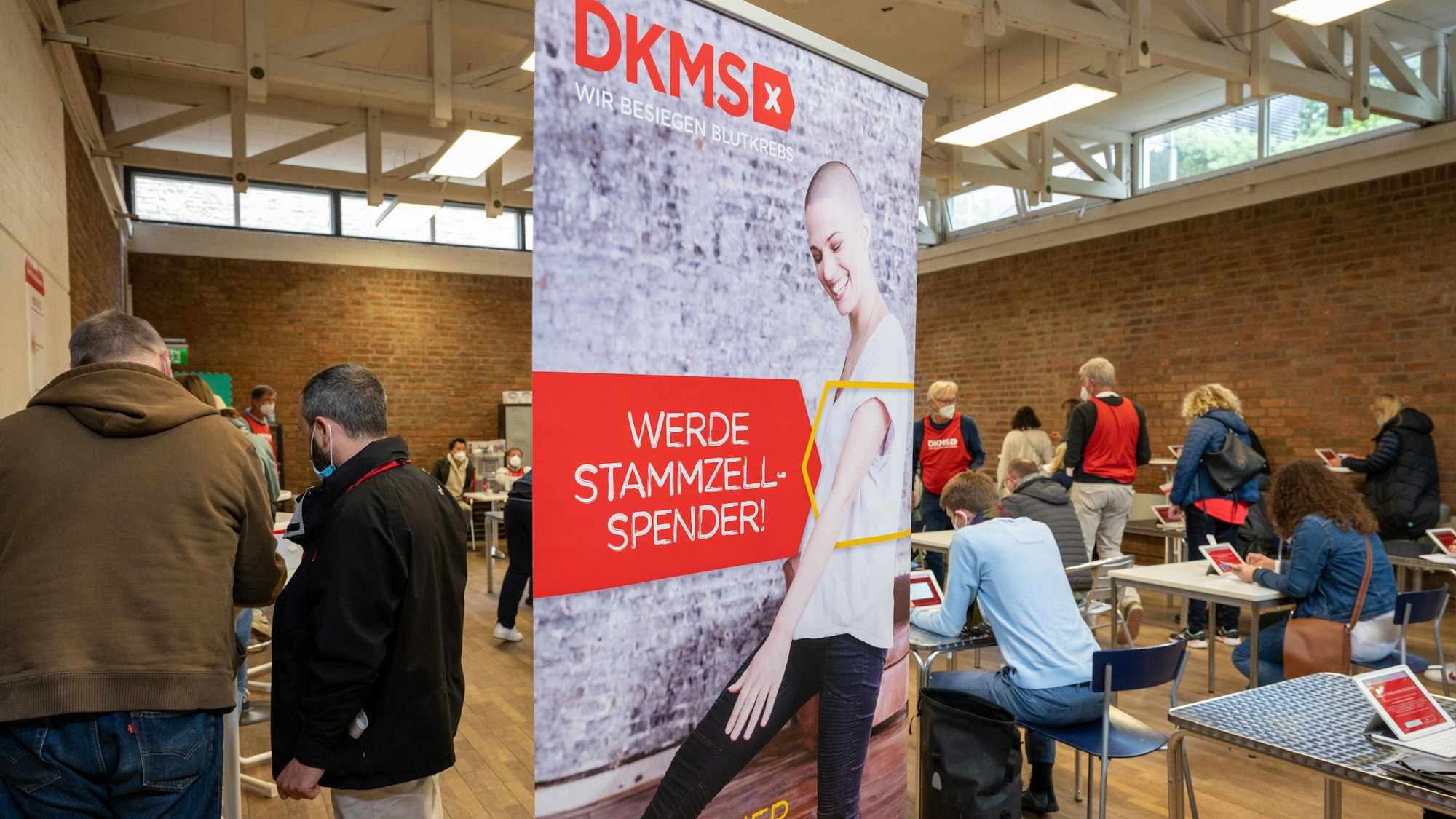 In einer Halle steht ein Plakat mit der Aufschrift „DKMS - Werde Stammzellspender“, daneben stehen und sitzen mehrere Menschen.
