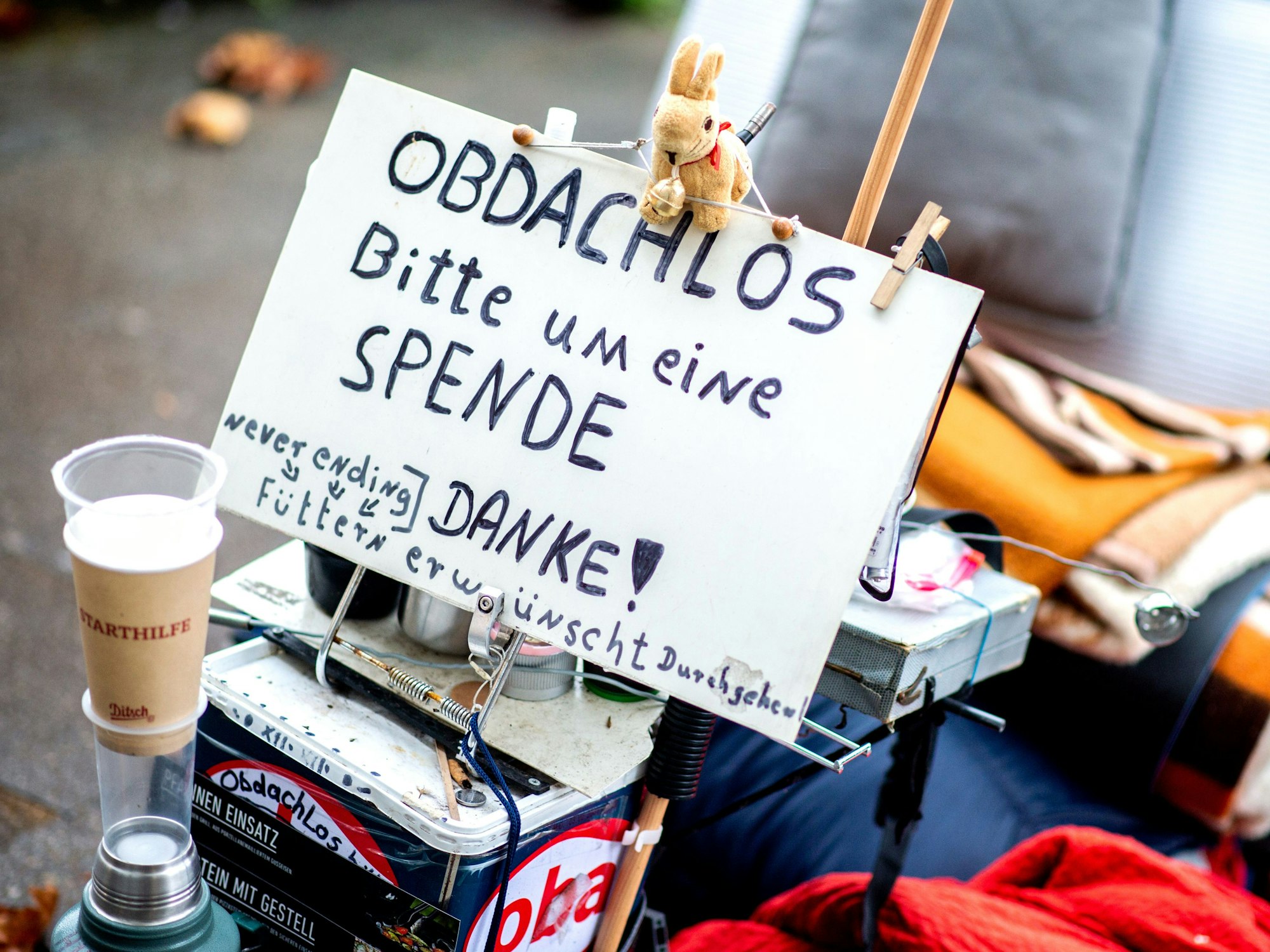 Ein Schild mit der Aufschrift „Obdachlos - bitte um eine Spende - danke!“ steht neben den Habseligkeiten eines Obdachlosen, die auf einem Bürgersteig in der Innenstadt liegen.