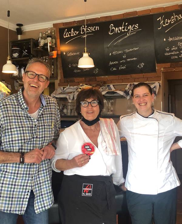 Besitzer Olaf Schlien, seine Mutter Brigitte Breiter und Köchin Esther Radermacher stehen vor der Theke und halten den Jode Lade-Aufkleber in die Kamera