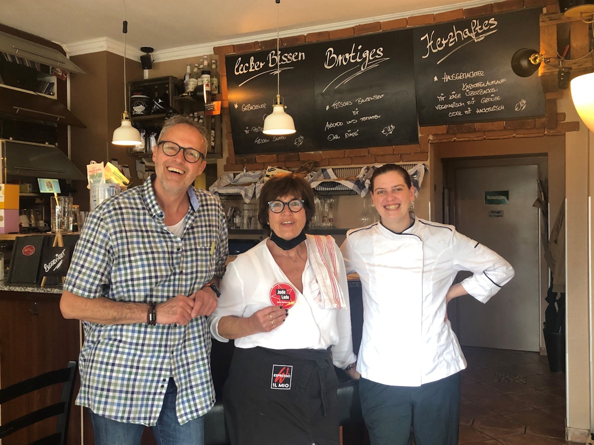 Besitzer Olaf Schlien, seine Mutter Brigitte Breiter und Köchin Esther Radermacher stehen vor der Theke und halten den Jode Lade-Aufkleber in die Kamera