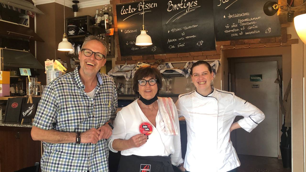 Besitzer Olaf Schlien, seine Mutter Brigitte Breiter und Köchin Esther Radermacher stehen vor der Theke und halten den Jode Lade-Aufkleber in die Kamera