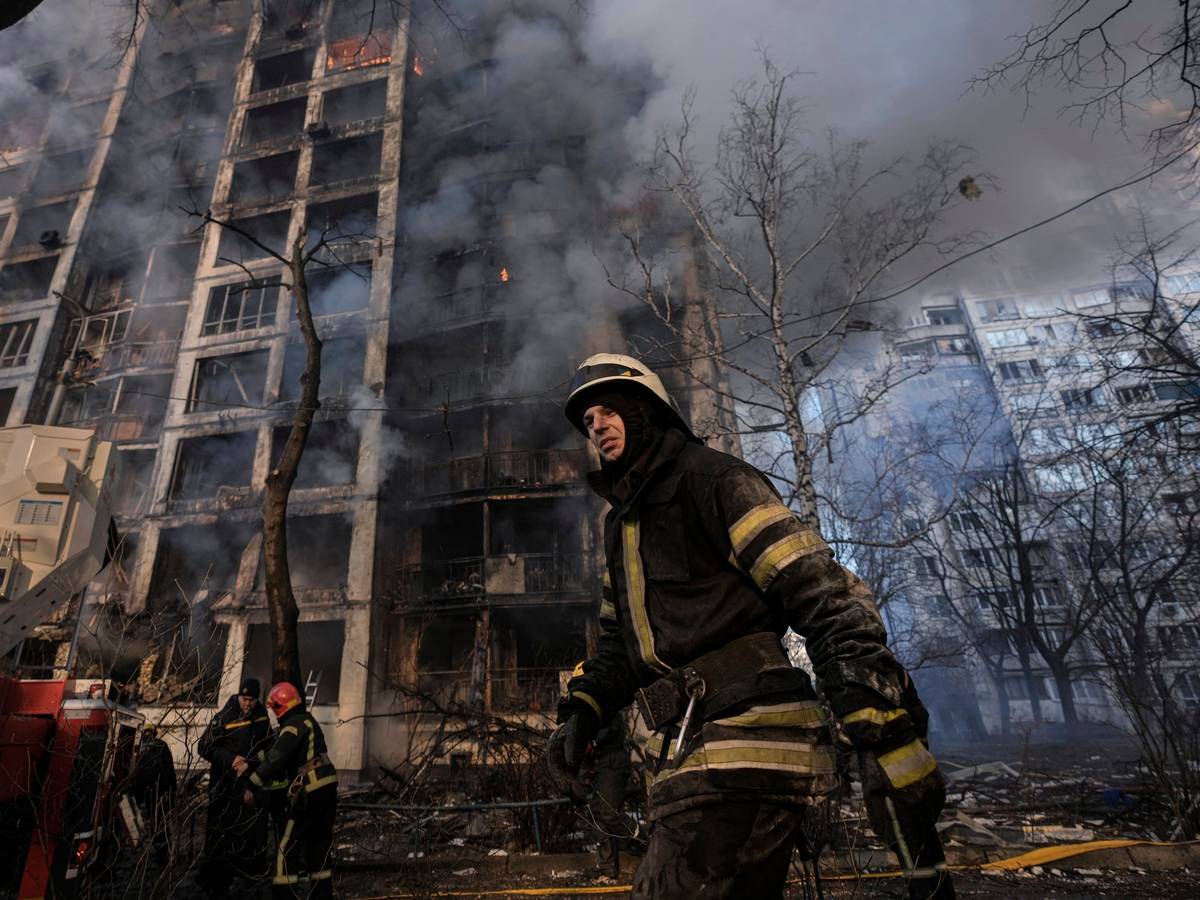 15.03.2022, Ukraine, Kyjiw: Einsatzkräfte der Feuerwehr stehen vor einem brennenden Wohngebäude, das durch Bombenbeschuss des russischen Militärs zerstört wurde.