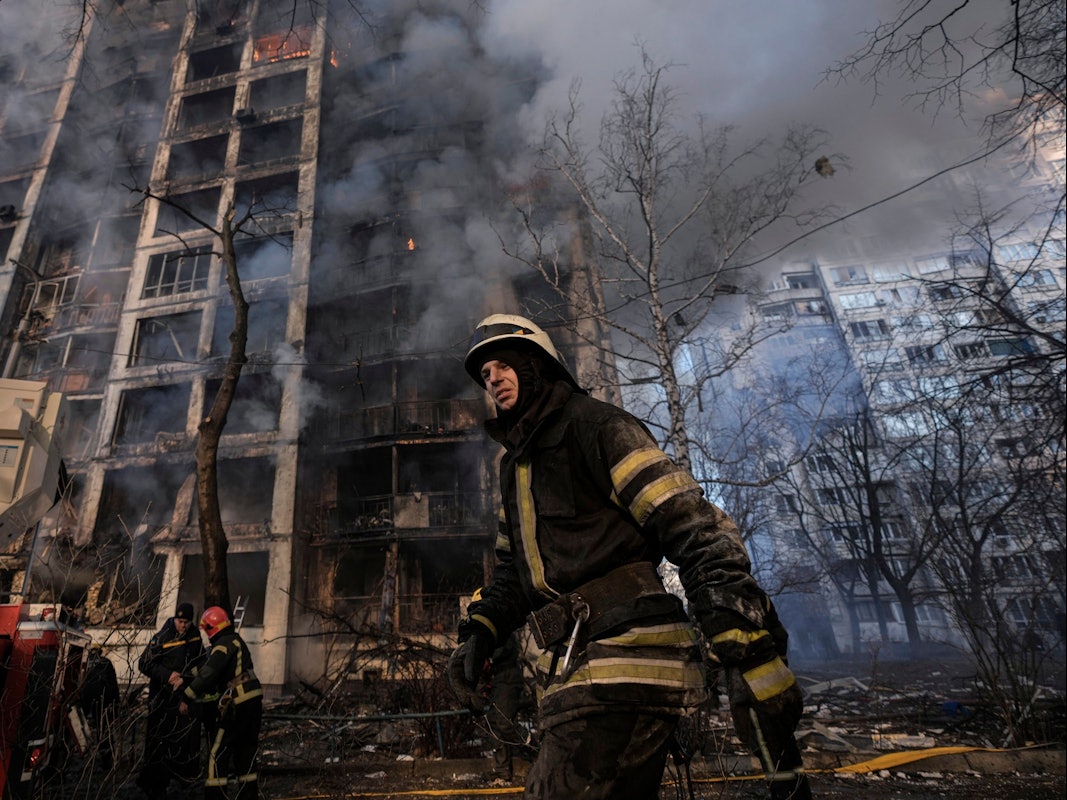 15.03.2022, Ukraine, Kyjiw: Einsatzkräfte der Feuerwehr stehen vor einem brennenden Wohngebäude, das durch Bombenbeschuss des russischen Militärs zerstört wurde.