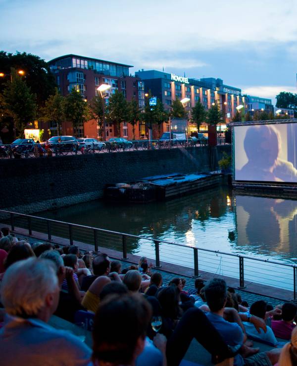 Menschen schauen sich im Open Air Kino am Rheinauhafen in Köln vor der Domkulisse einen Film an.