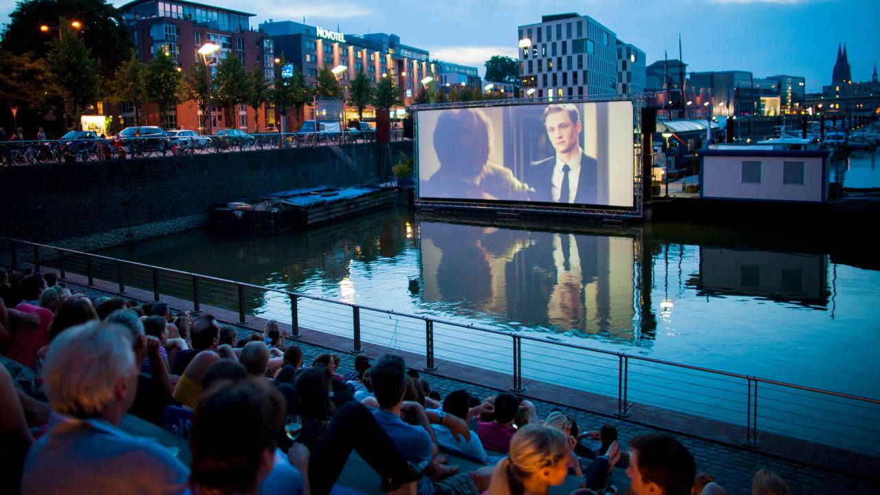Menschen schauen sich im Open Air Kino am Rheinauhafen in Köln vor der Domkulisse einen Film an.