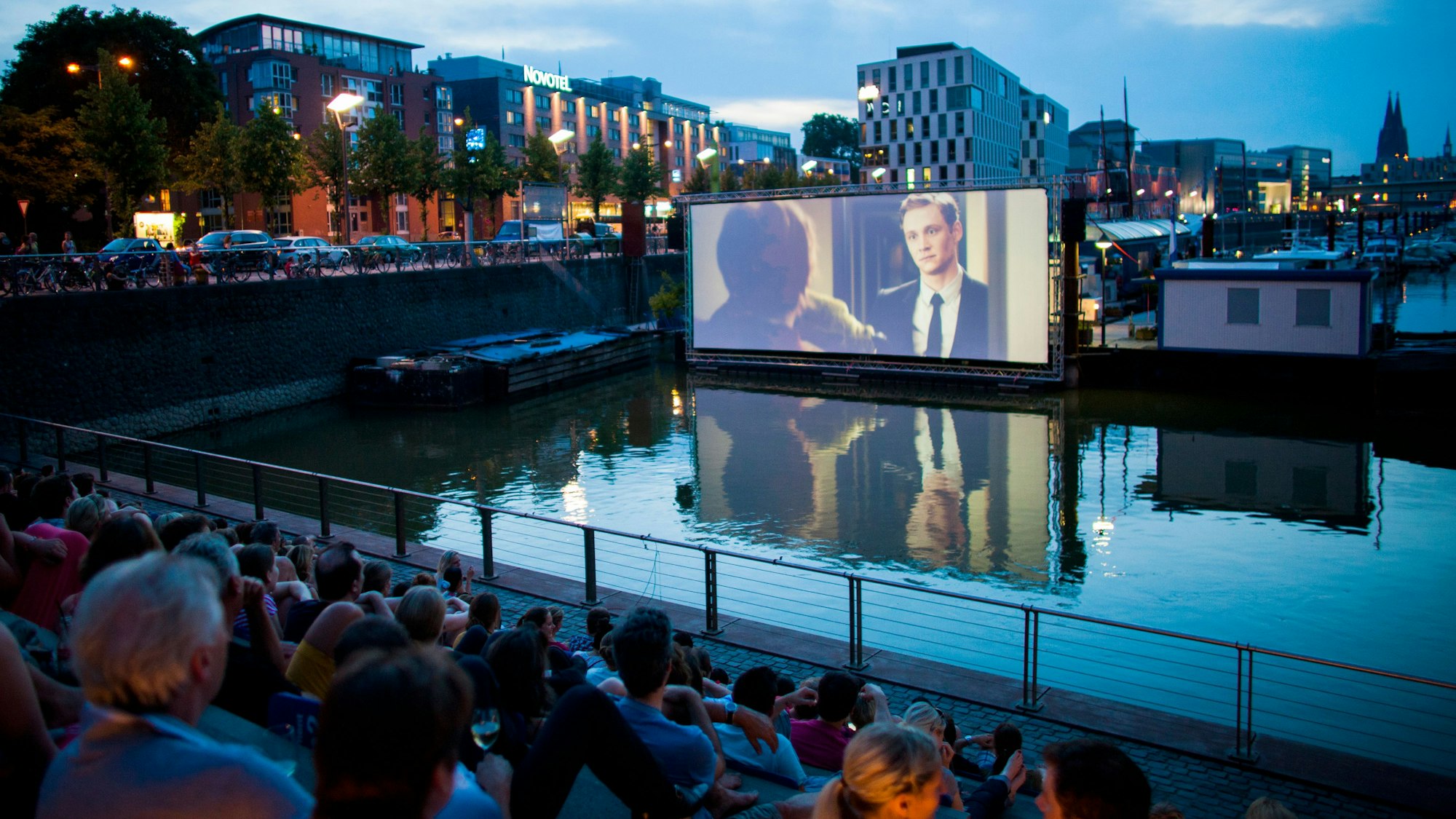 Menschen schauen sich im Open Air Kino am Rheinauhafen in Köln vor der Domkulisse einen Film an.