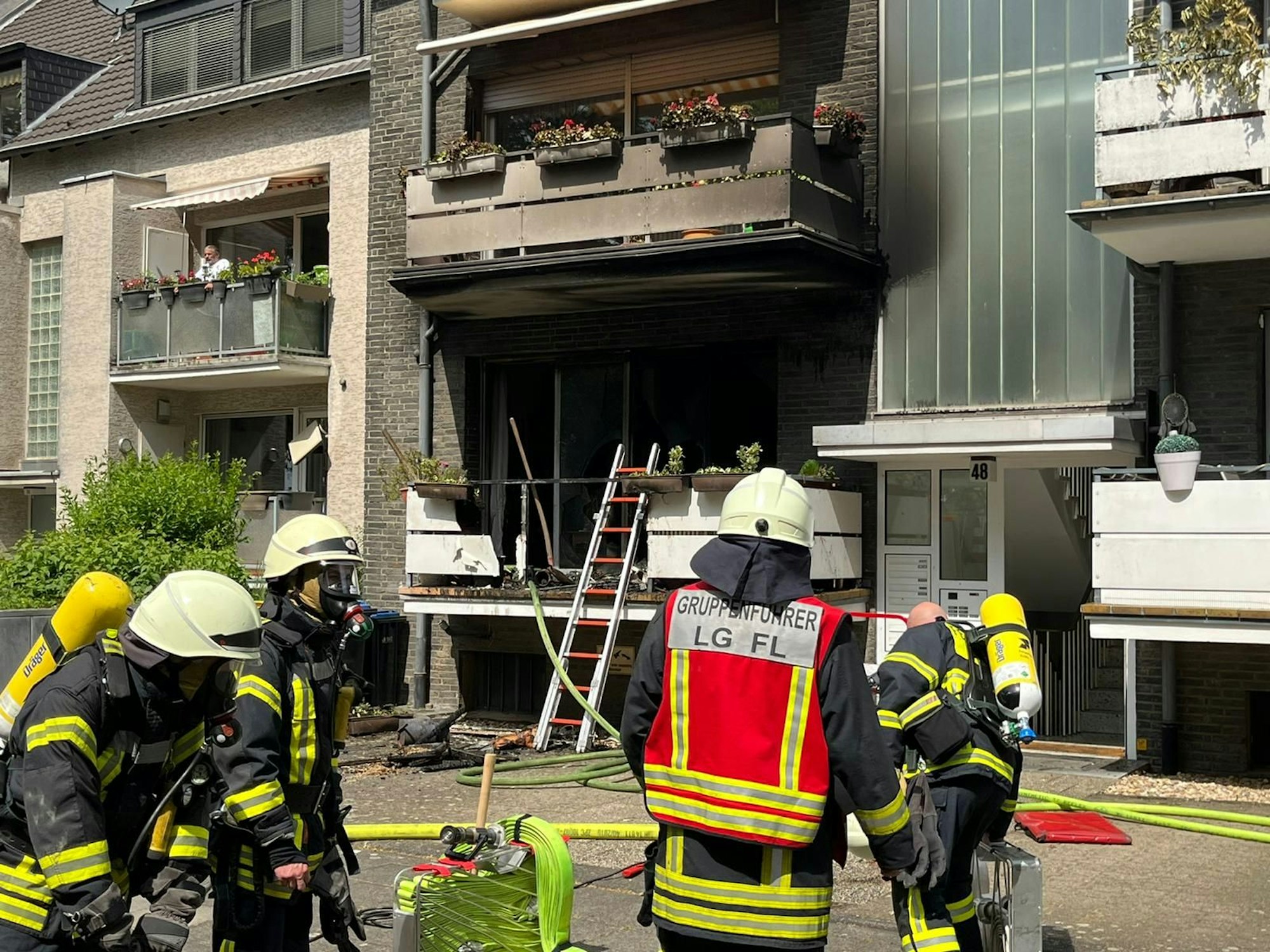 Mehrere Feuerwehrmänner befinden sich vor einem Mehrfamilienhaus. Im Parterre ist ein Balkon pechschwarz vor Ruß.