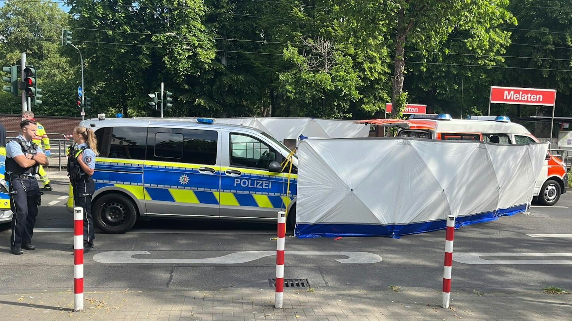 Einsatzfahrzeuge und -kräfte stehen auf der Straße vor der KVB-Haltestelle Melaten. Ein Sichtschutz ist aufgebaut.
