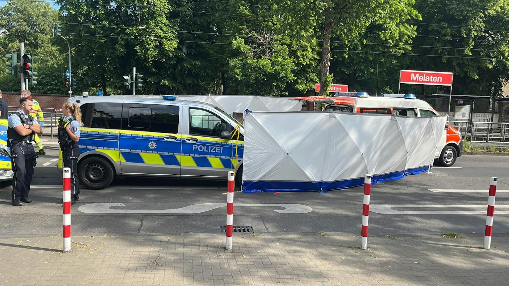 Auf der Straße ist ein Blickschutz aufgebaut, daneben stehen eine Polizistin und ein Polizist neben einem Polizeifahrzeug. Im Hintergrund erkennt man ein Schild mit der Aufschrift „Melaten“.
