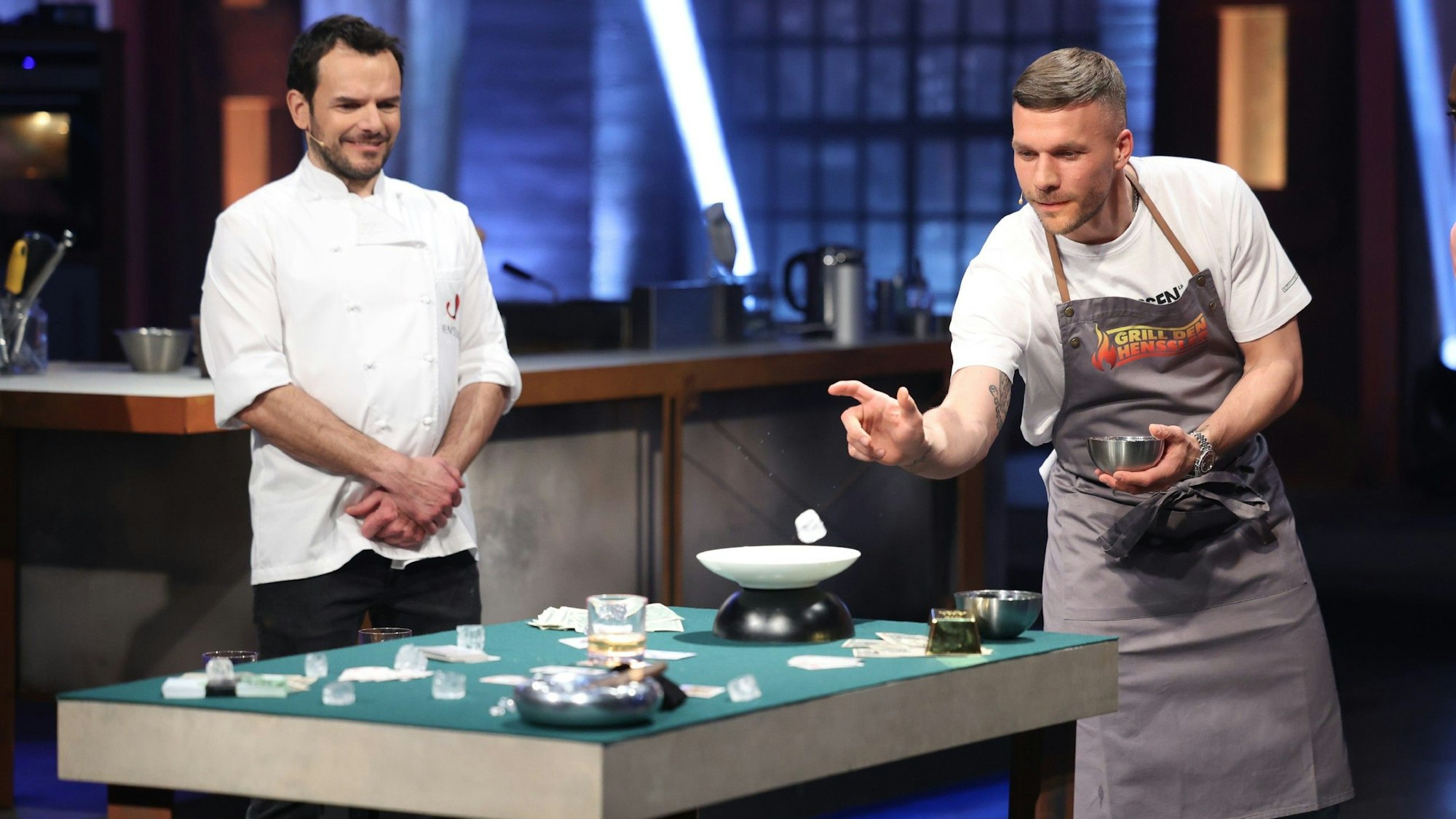 Lukas Podolski (r.) versucht, Eiswürfel in ein Whiskey-Glas zu werfen. Daneben steht Steffen Henssler (l.) in der Vox-Show „Grill den Henssler“.