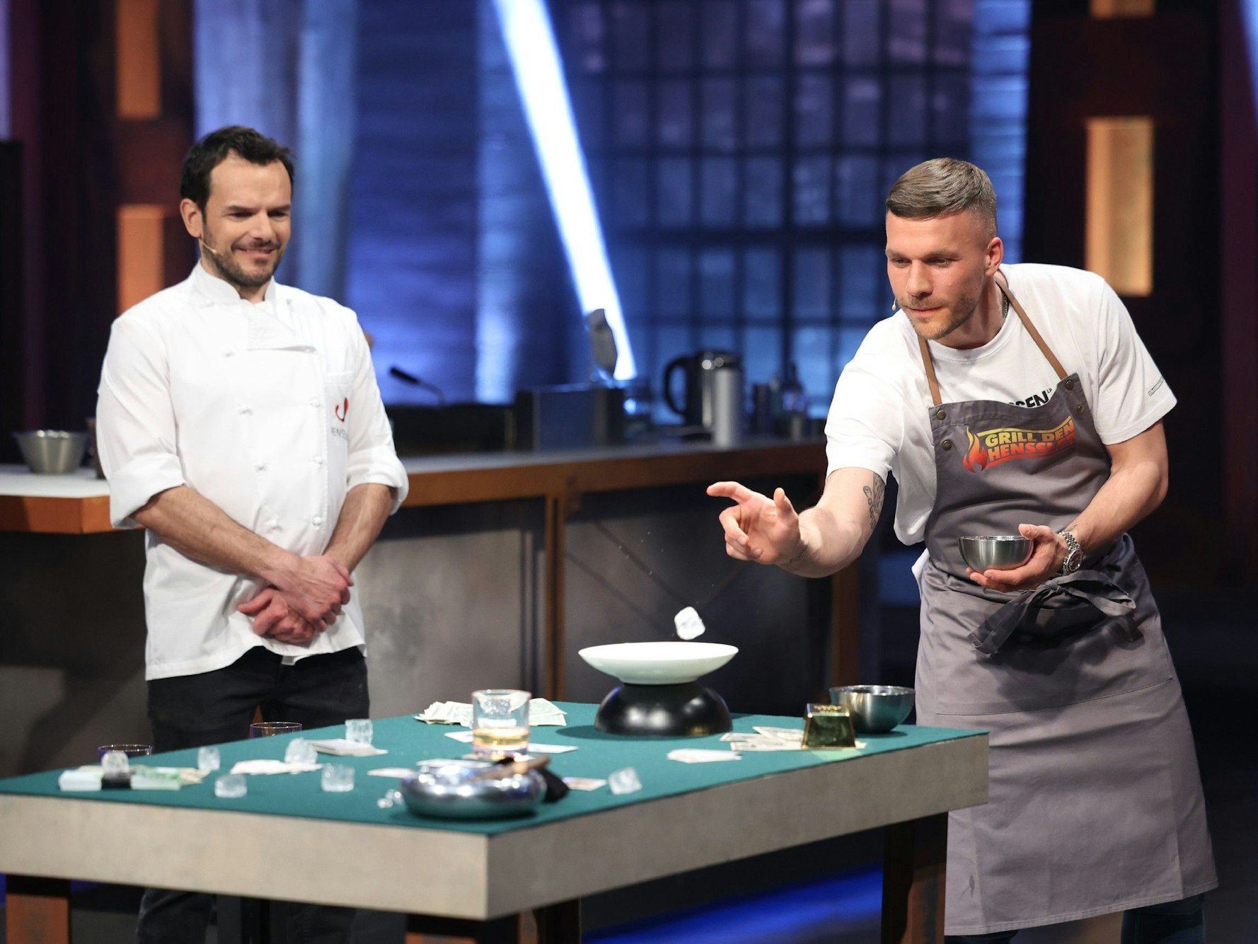 Lucas Podolski (r.) versucht, Eiswürfel in ein Whiskey-Glas zu werfen. Daneben steht Steffen Henssler (l.) in der Vox-Show „Grill den Henssler“ am 15. Mai 2022.
RTL/Frank Hempel