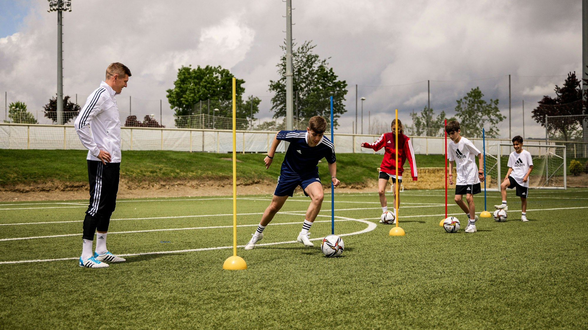 Toni Kroos beobachtet Kinder, die um Stangen dribbeln.
