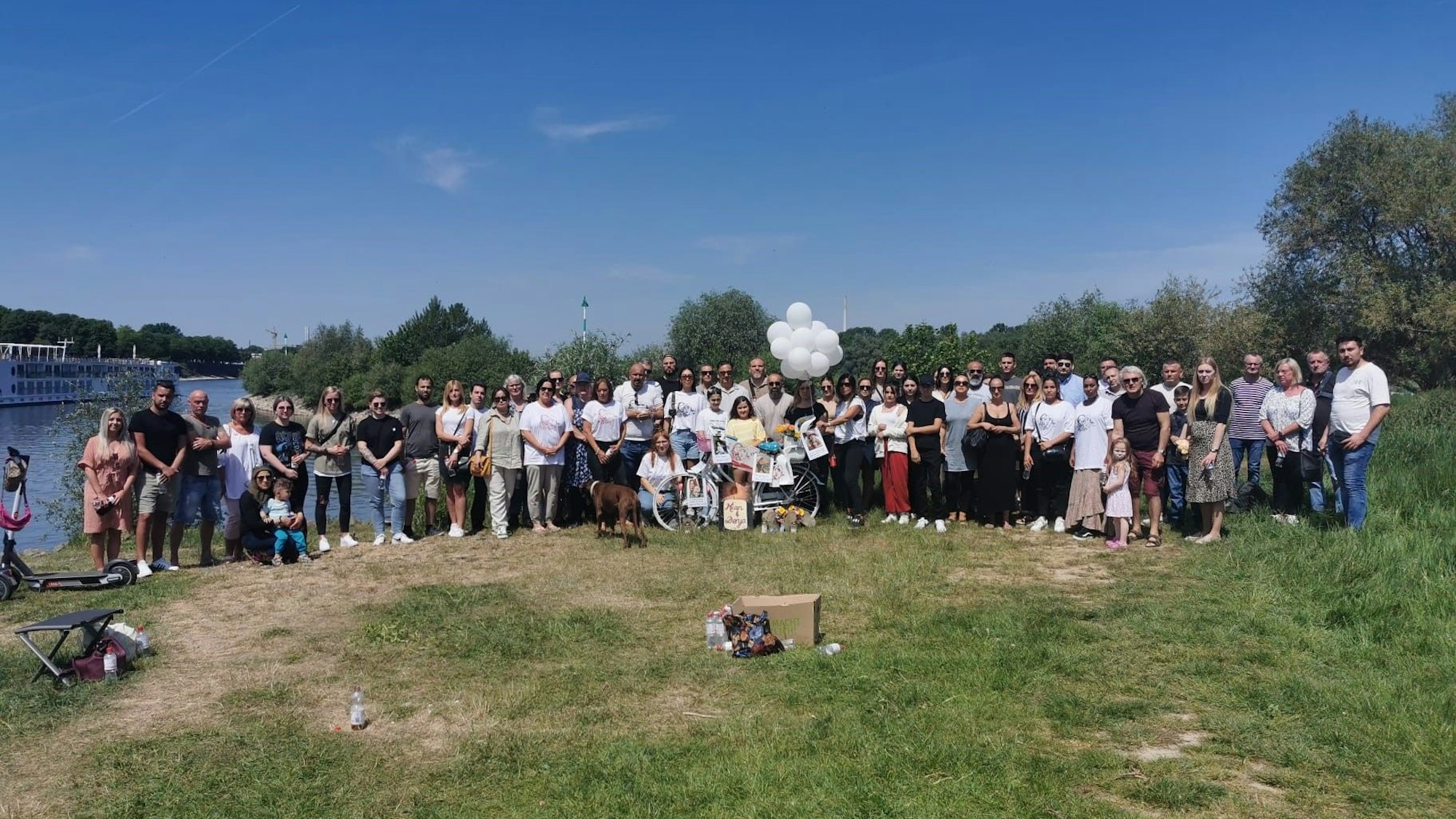 Eine Menschengruppe steht auf einer Wiese am Rhein.