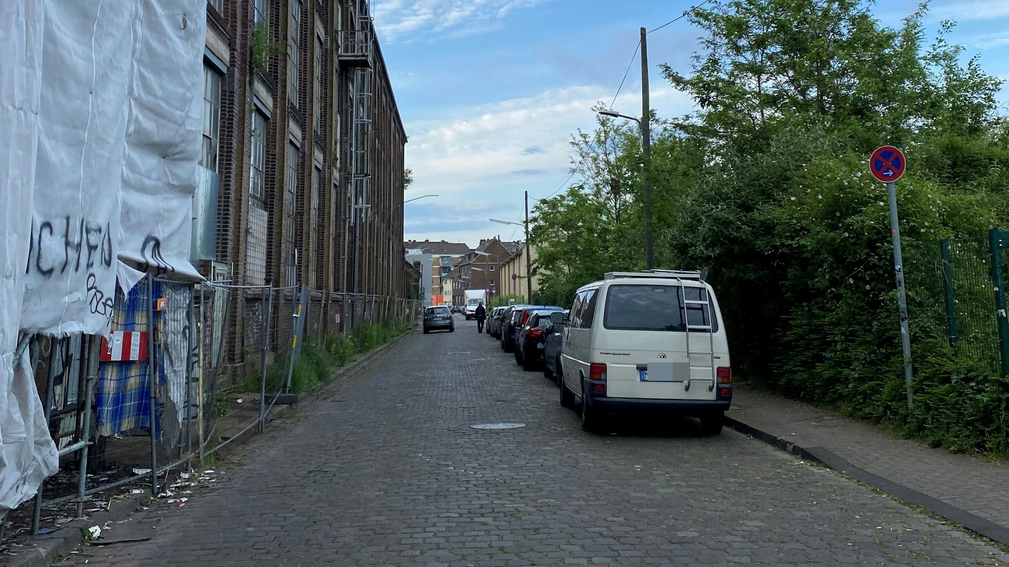 Blick in die Neuerburgstraße in Köln-Kalk: Rechts stehen parkende Autos, links befindet sich ein langgestrecktes Gebäude, an dem sich zum Teil ein Gerüst mit Planen befindet.
