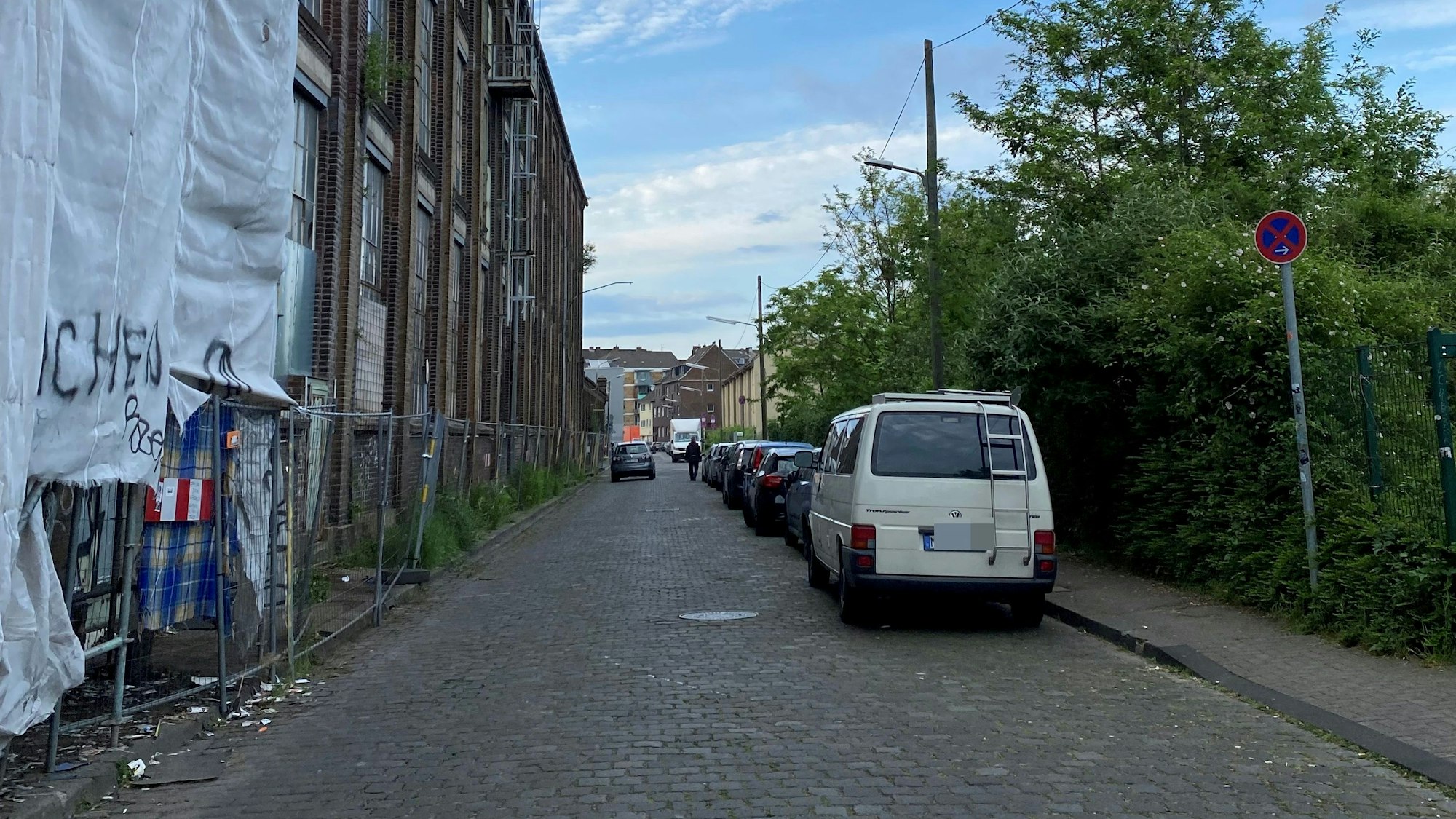 Blick in die Neuerburgstraße in Köln-Kalk: Rechts stehen parkende Autos, links befindet sich ein langgestrecktes Gebäude, an dem sich zum Teil ein Gerüst mit Planen befindet.