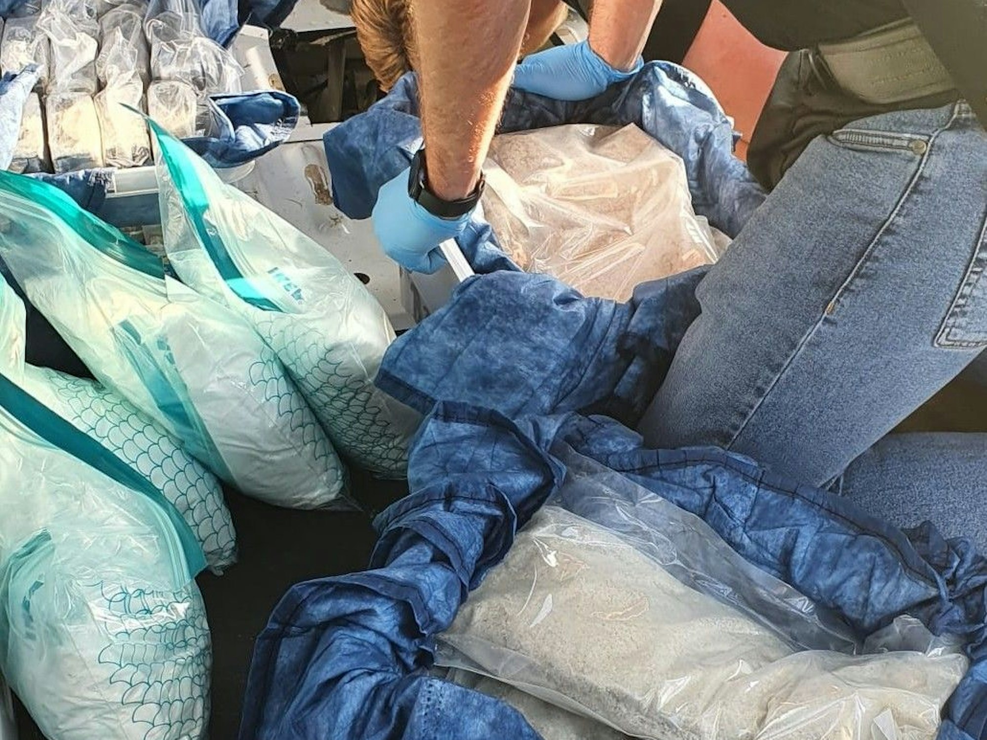 Ein Mann greift nach einem Behälter, in dem eine Tüte mit weißem Pulver ist. Um ihn herum stehen weitere Behälter sowie Tüten mit weißem Pulver beziehungsweise Pillen.