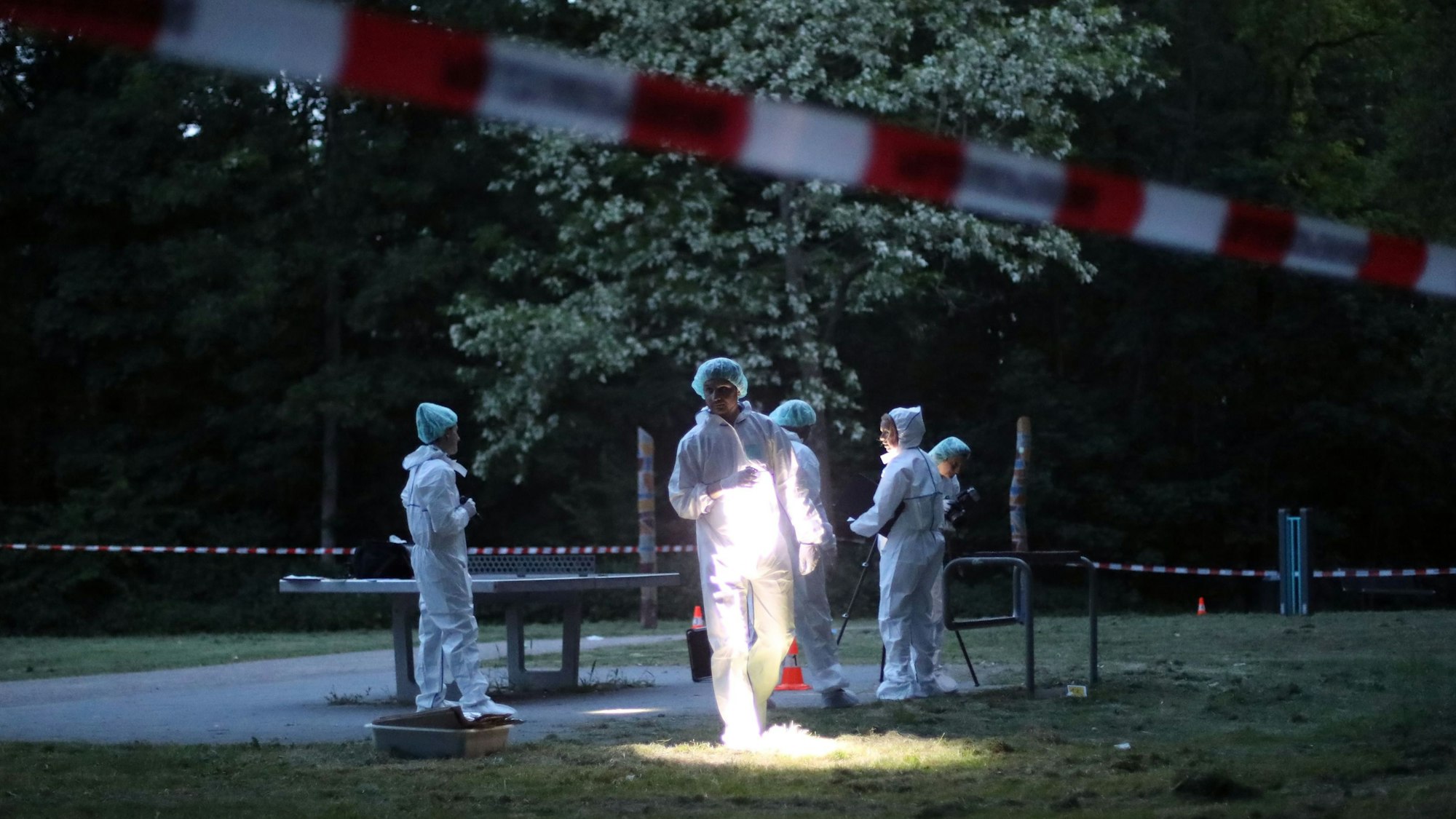 Kräfte der Spurensicherung in Schutzanzügen stehen an einer Polizeiabsperrung nahe einer Tischtennisplatte, ein Beamter leuchtet mit einer Lampe auf den Rasen.