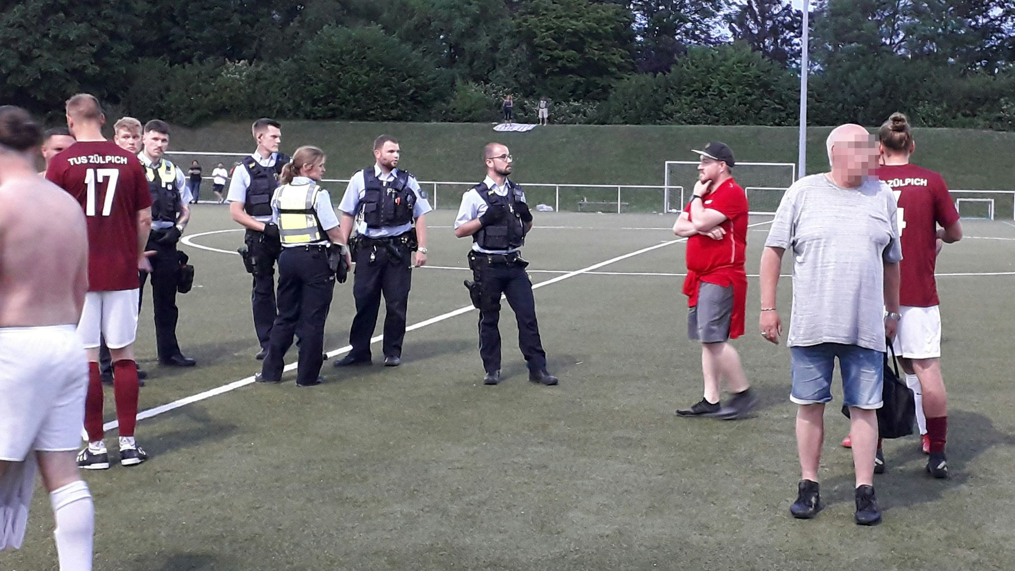 Polizeikräfte stehen mit Spielern und Trainern auf einem Fußballplatz.