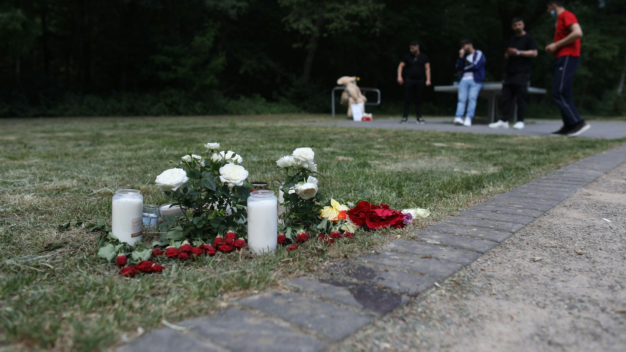 Auf einem Spielplatz liegen Blumen und stehen Kerzen. Im Hintergrund befinden sich mehrere Jugendliche an einer Tischtennisplatte.
