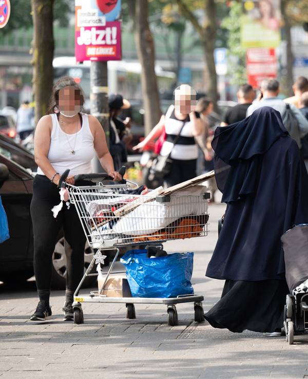 Eine Frau steht mit einem Einkaufswagen auf einem Gehweg in Köln.