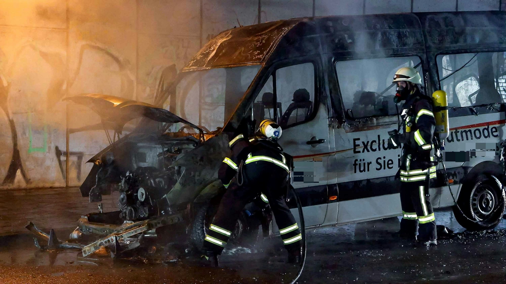 Zwei Feuerwehrmänner stehen an einem ausgebrannten Kleintransporter.