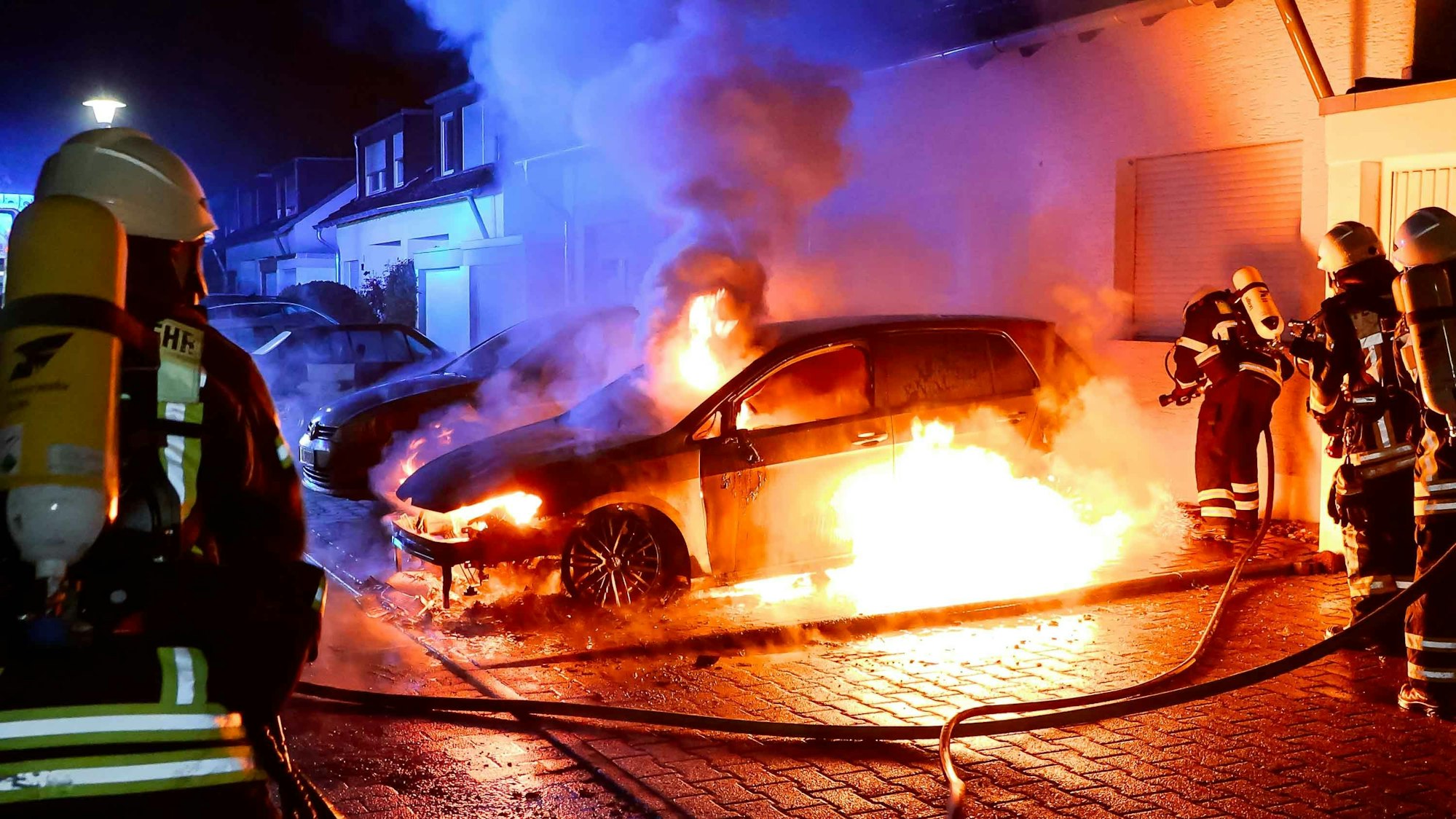 Ein Auto brennt lichterloh vor einem Haus, mehrere Feuerwehrmänner sind im Einsatz.