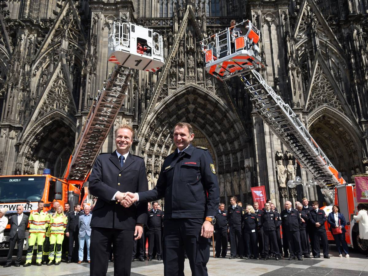 Kräfte sowie Einsatzfahrzeuge der Feuerwehren Köln und Düsseldorf präsentieren sich auf der Domplatte, im Vordergrund schütteln sich die Chefs die Hand.