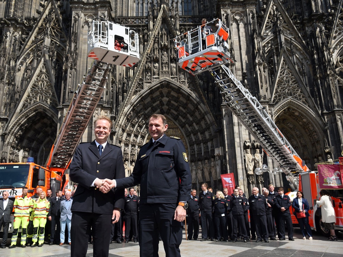 Kräfte sowie Einsatzfahrzeuge der Feuerwehren Köln und Düsseldorf präsentieren sich auf der Domplatte, im Vordergrund schütteln sich die Chefs die Hand.