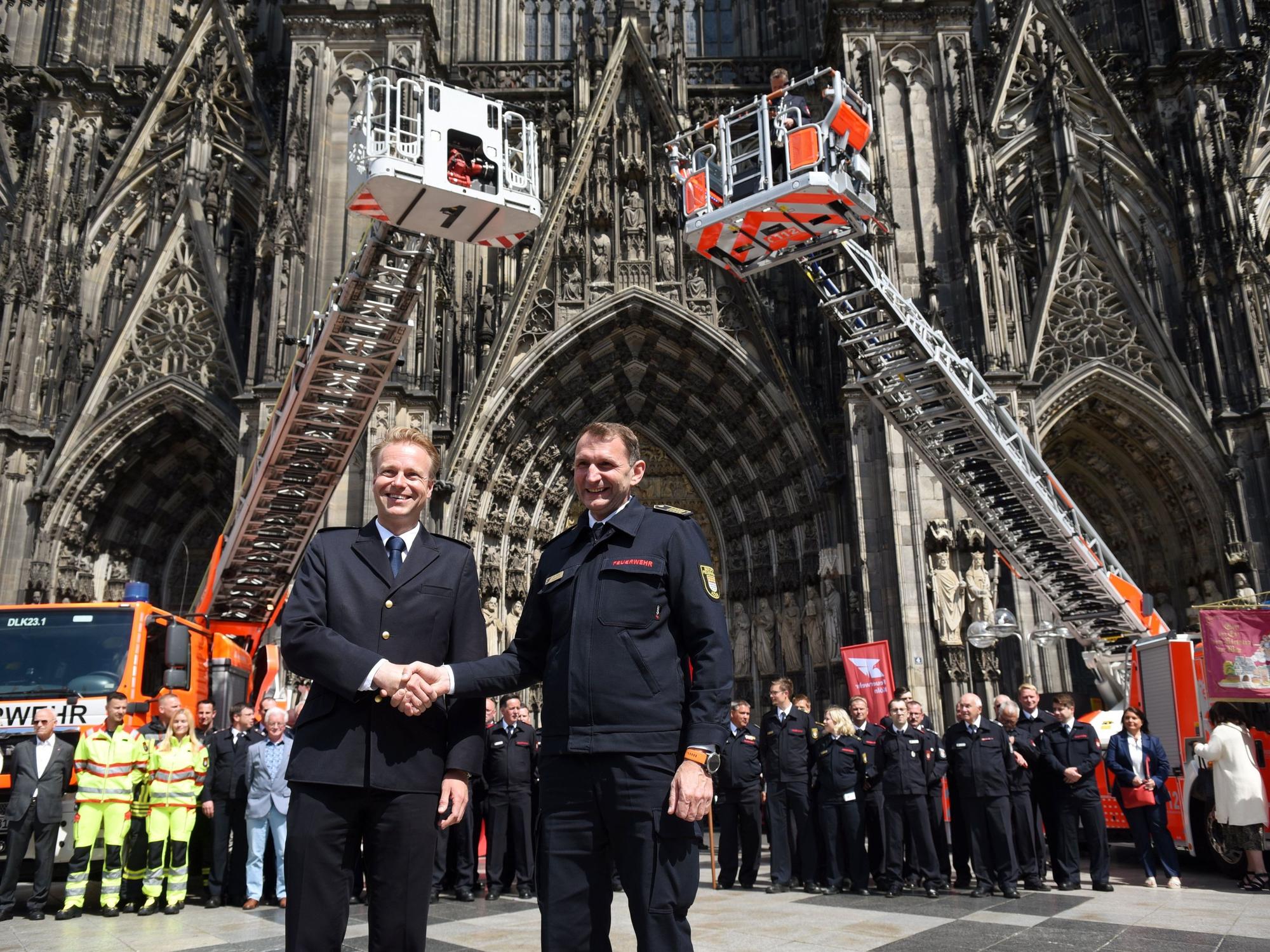 Kräfte sowie Einsatzfahrzeuge der Feuerwehren Köln und Düsseldorf präsentieren sich auf der Domplatte, im Vordergrund schütteln sich die Chefs die Hand.