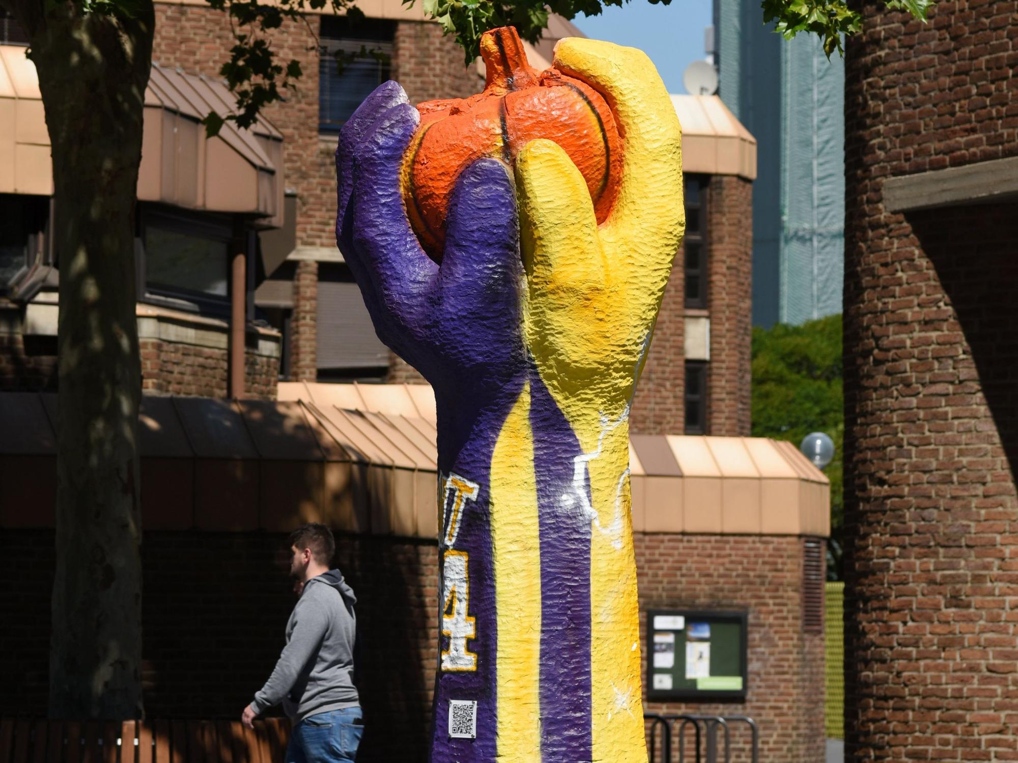 Die Skulptur „Tomate“ zeigt zwei Hände, die eine Tomate halten. Sie ist in den Farben gelb und lila bemalt und erinnert an das Trikot des verstorbenen Basketballstars Kobe Bryant.