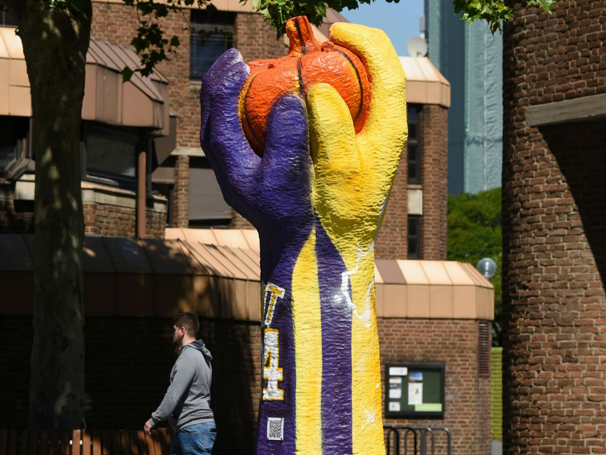 Die Skulptur „Tomate“ zeigt zwei Hände, die eine Tomate halten. Sie ist in den Farben gelb und lila bemalt und erinnert an das Trikot des verstorbenen Basketballstars Kobe Bryant.