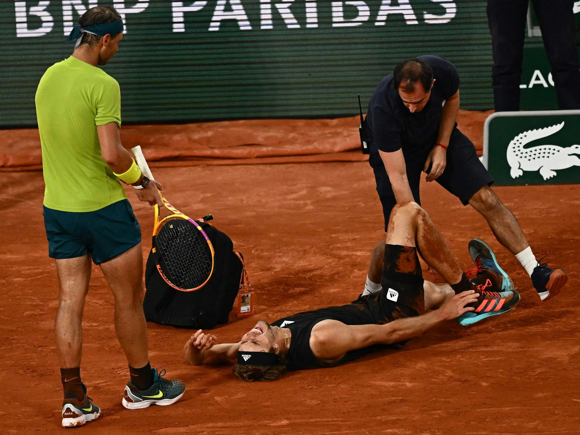 Alexander Zverev liegt schreiend am Boden, während ihn ein Arzt behandelt. Gegenspieler Rafael Nadal leidet daneben mit.