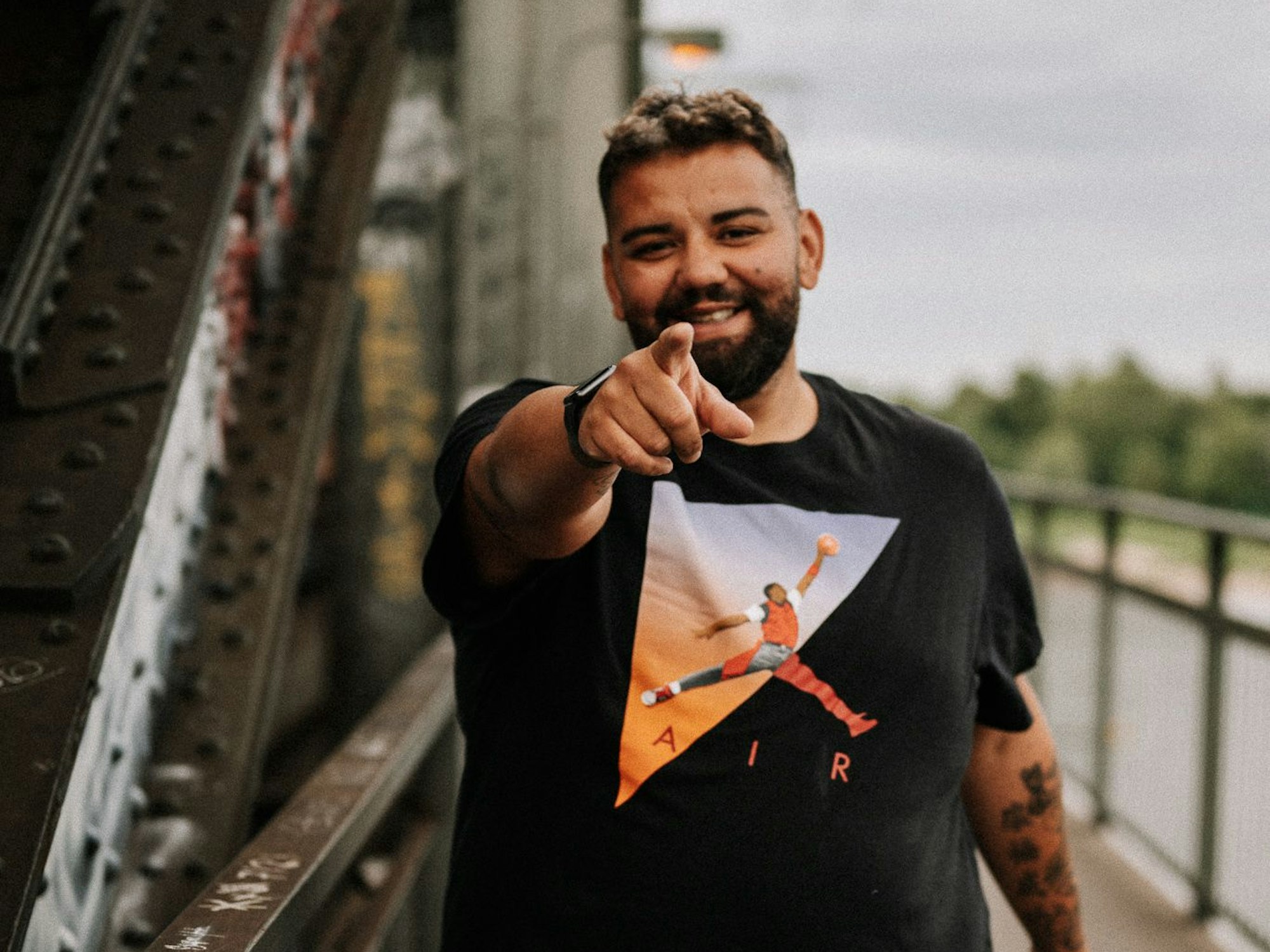 Marcello Volk geht über eine Brücke in Köln und zeigt auf die Kamera.