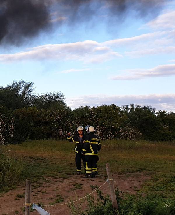Hinter einem Gebüsch steigt eine schwarze Rauchsäule in den Himmel. Ein paar Meter entfernt stehen zwei Feuerwehrmänner.