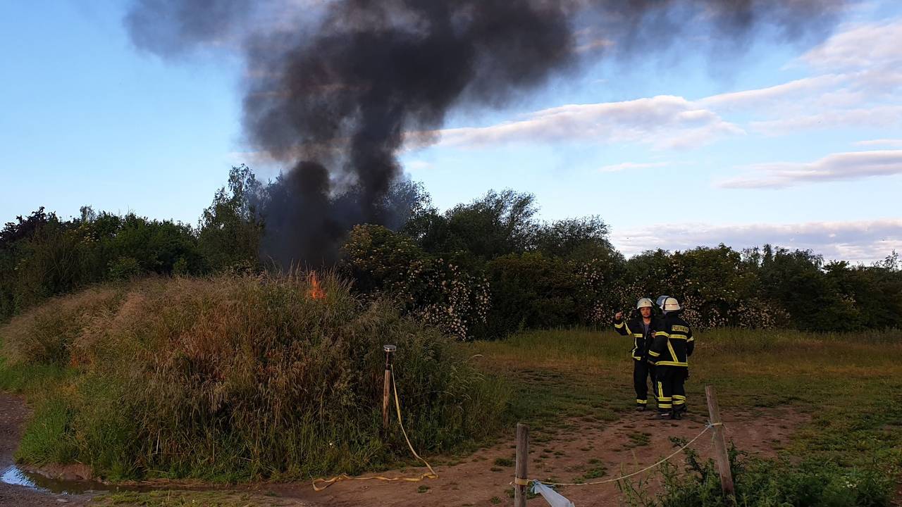 Hinter einem Gebüsch steigt eine schwarze Rauchsäule in den Himmel. Ein paar Meter entfernt stehen zwei Feuerwehrmänner.