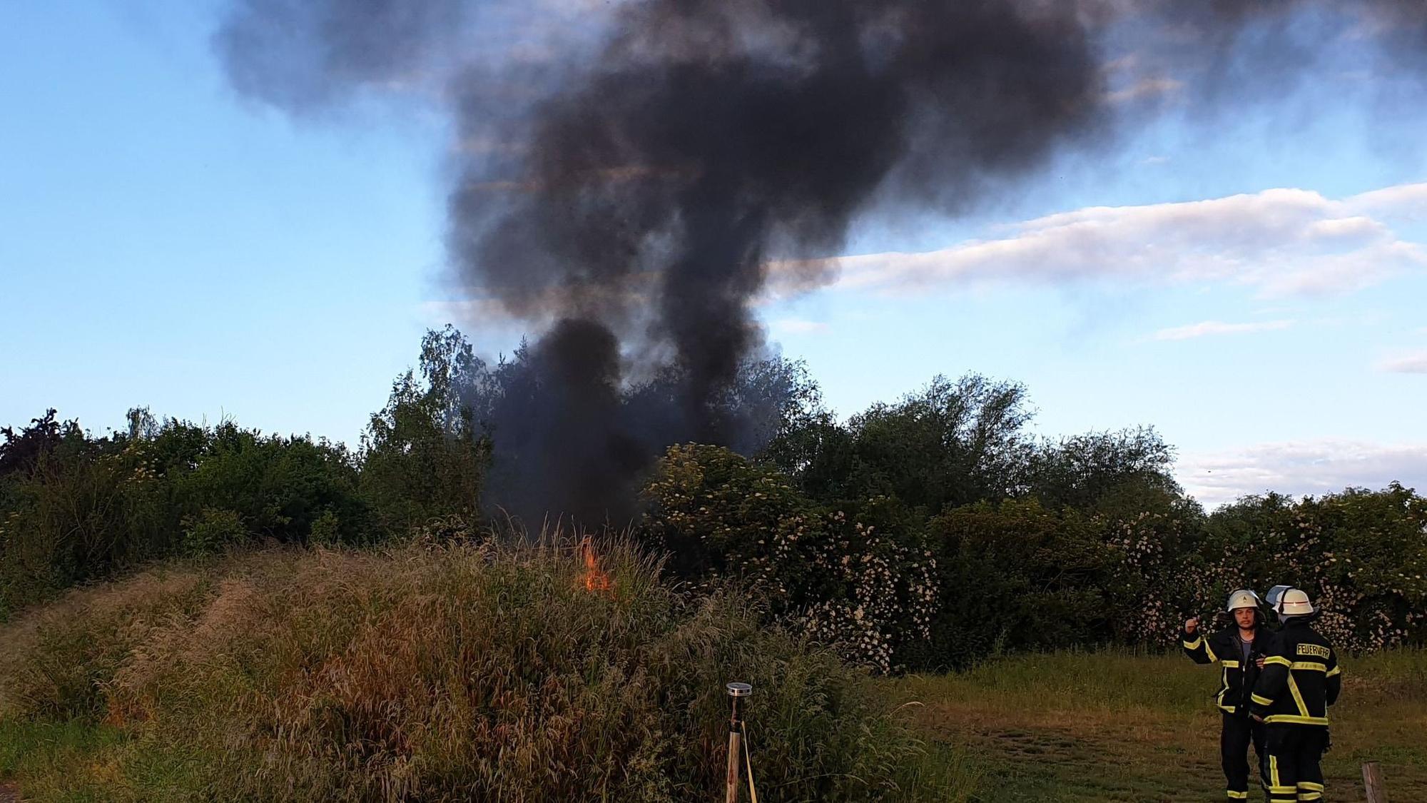 Hinter einem Gebüsch steigt eine schwarze Rauchsäule in den Himmel. Ein paar Meter entfernt stehen zwei Feuerwehrmänner.