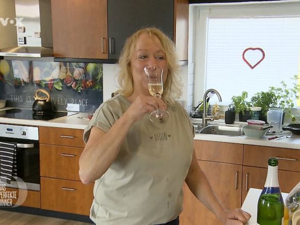 Friseurmeisterin Hiltrud steht bei „Das perfekte Dinner“ mit einem Glas Sekt in der Küche.
