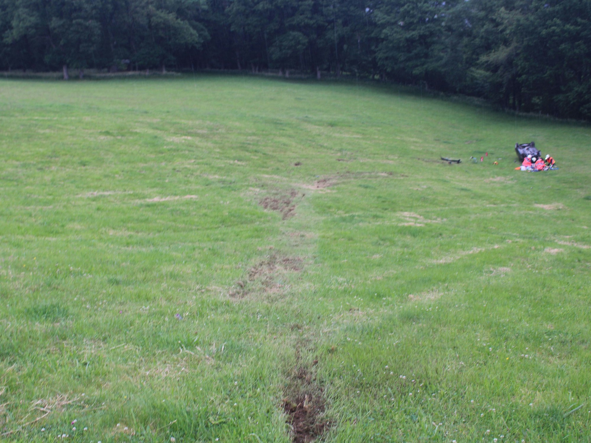Tiefe Spuren im Wiesenhang, an dessen Ende ein Auto auf dem Dach liegt, Rettungskräfte knien um den Fahrer.