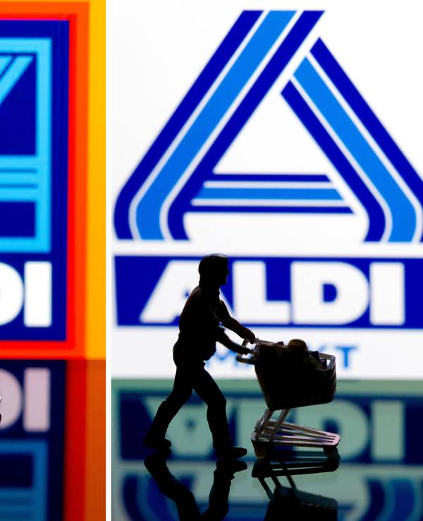 Eine kleine Plastikfigur mit einem Einkaufswagen in Berlin vor einem Logo von Aldi Süd (links) und Aldi Nord