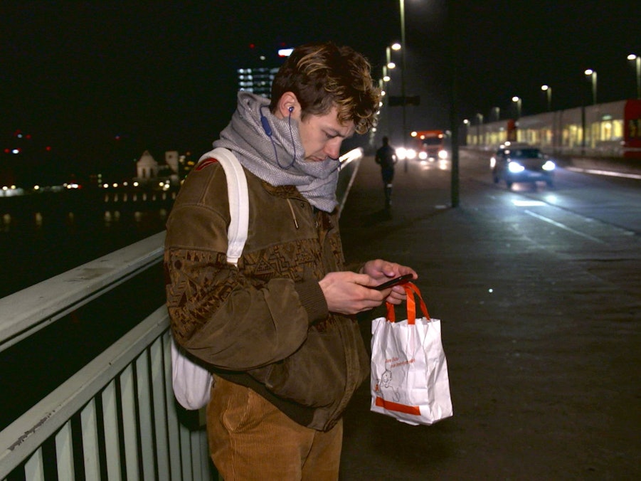 Schauspieler Tim-Fabian Hoffmann steht Nachts auf der Deutzer Brücke und schaut auf sein Handy.